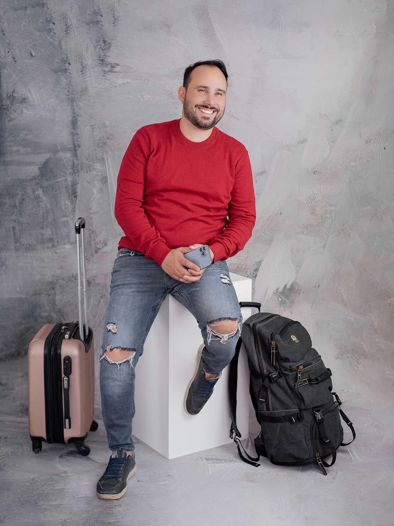 Zé Roberto sentado, de suéter vermelho, com um celular na mão, uma mala de viagem de um lado e uma mochila do outro